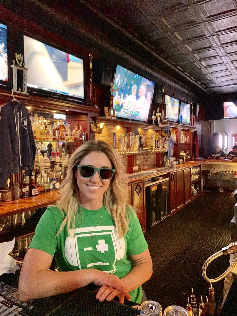 Woman with sunglasses and green shirt at a bar, smiling. Background features bar, TVs, alcohol bottles.
