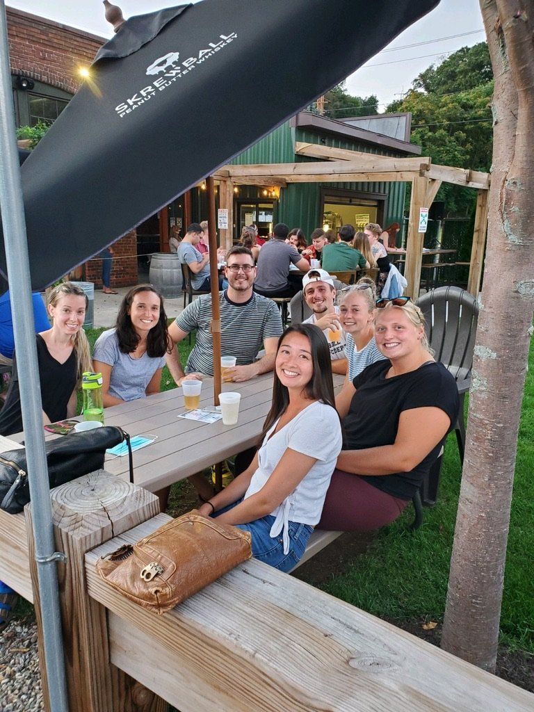Group of people at an outdoor table. Some are smiling, drinking, and looking at the camera. A bar is visible in the background.