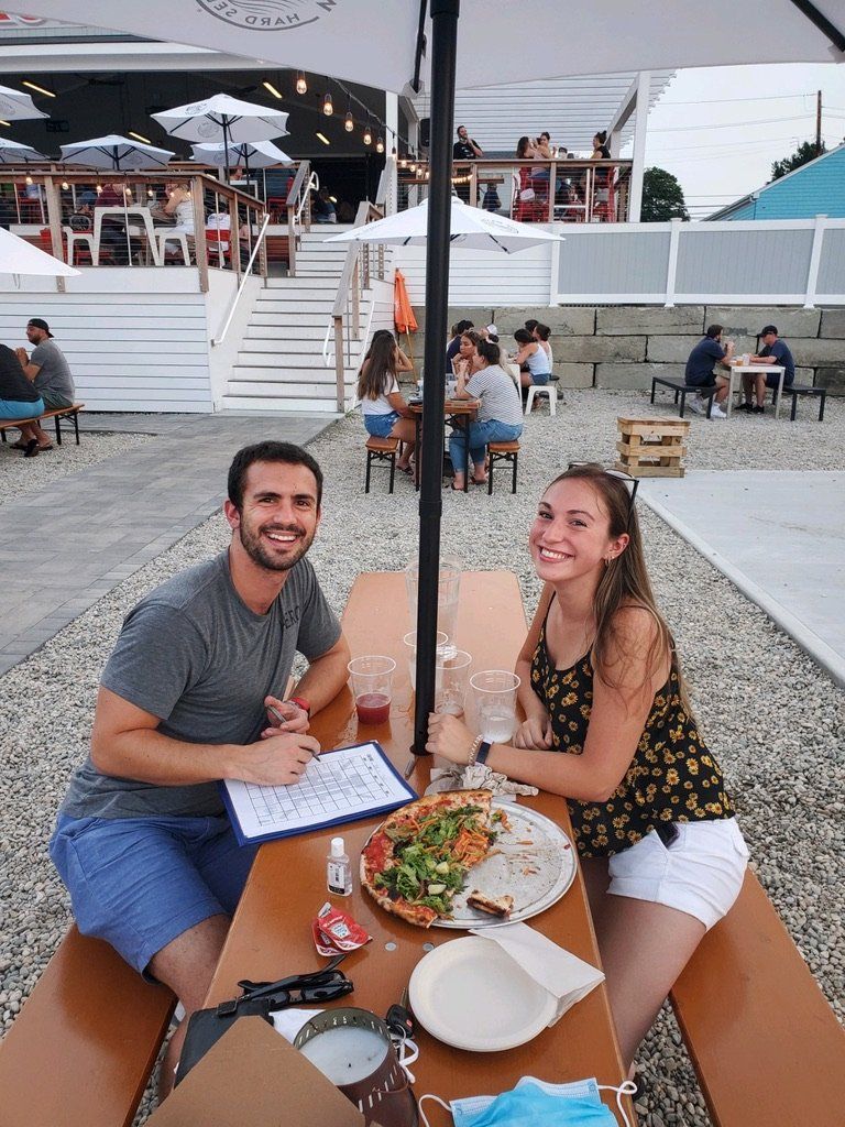 Couple at outdoor table with pizza and drinks, smiling. Restaurant setting with umbrellas.