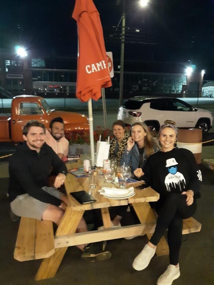 Group of people at a picnic table outdoors at night; orange umbrella, truck, and cars in background.