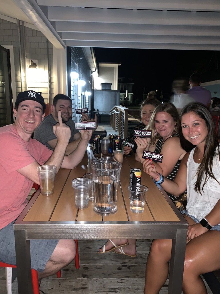 A group of friends at an outdoor bar, smiling, holding promotional signs, and drinking at a table.