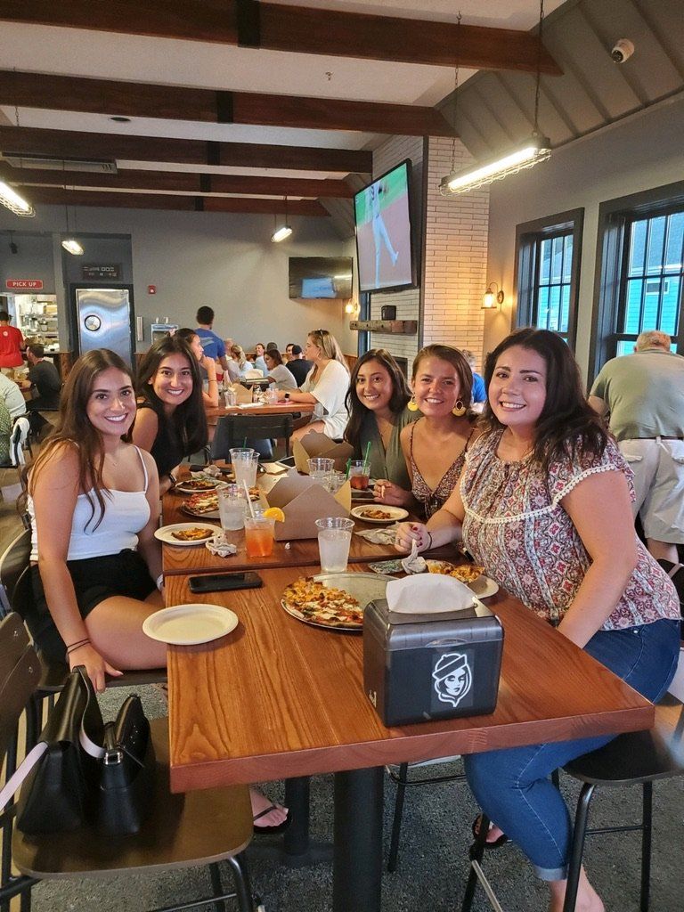 Group of friends at a restaurant, smiling, eating pizza. Wooden table, casual setting, indoor, daytime.