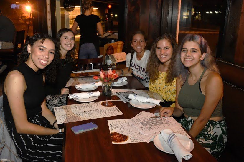 Six people at a restaurant table, smiling. Menus and plates are on the table. Dim lighting.