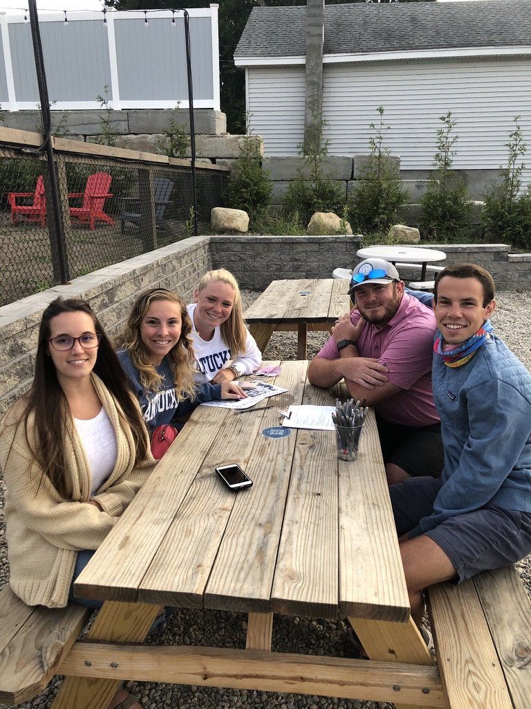 Group of people sitting at a picnic table outdoors, smiling. Table has papers and a phone. Buildings in background.