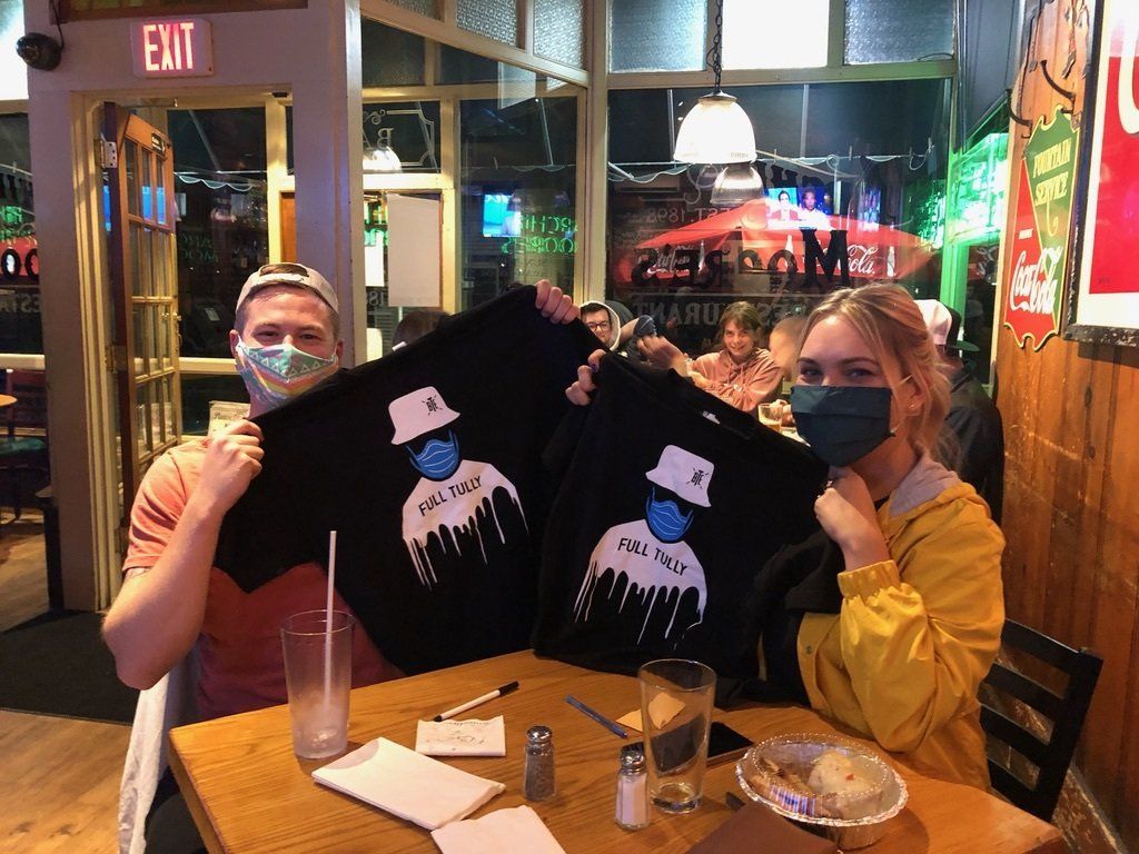Two masked people in a restaurant hold up black t-shirts with cartoon designs; other diners in background.