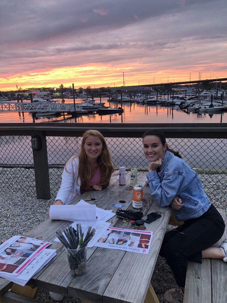 Two people at a wooden table with papers, pens, and drinks, harbor view, sunset.
