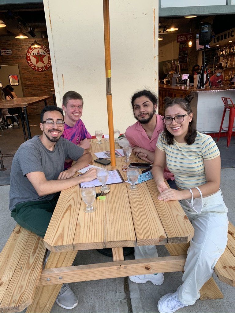 Four people seated at a wooden picnic table under an umbrella outside a restaurant, smiling.