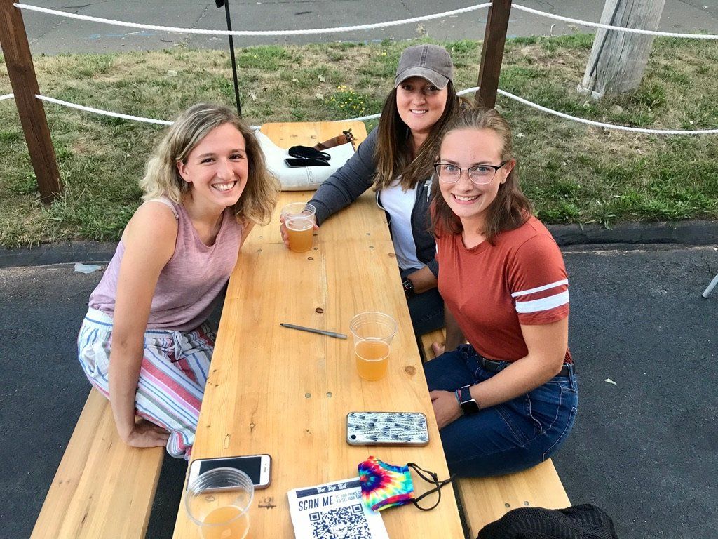 Three smiling women at a wooden picnic table, each with a beer. Outdoors, daytime.