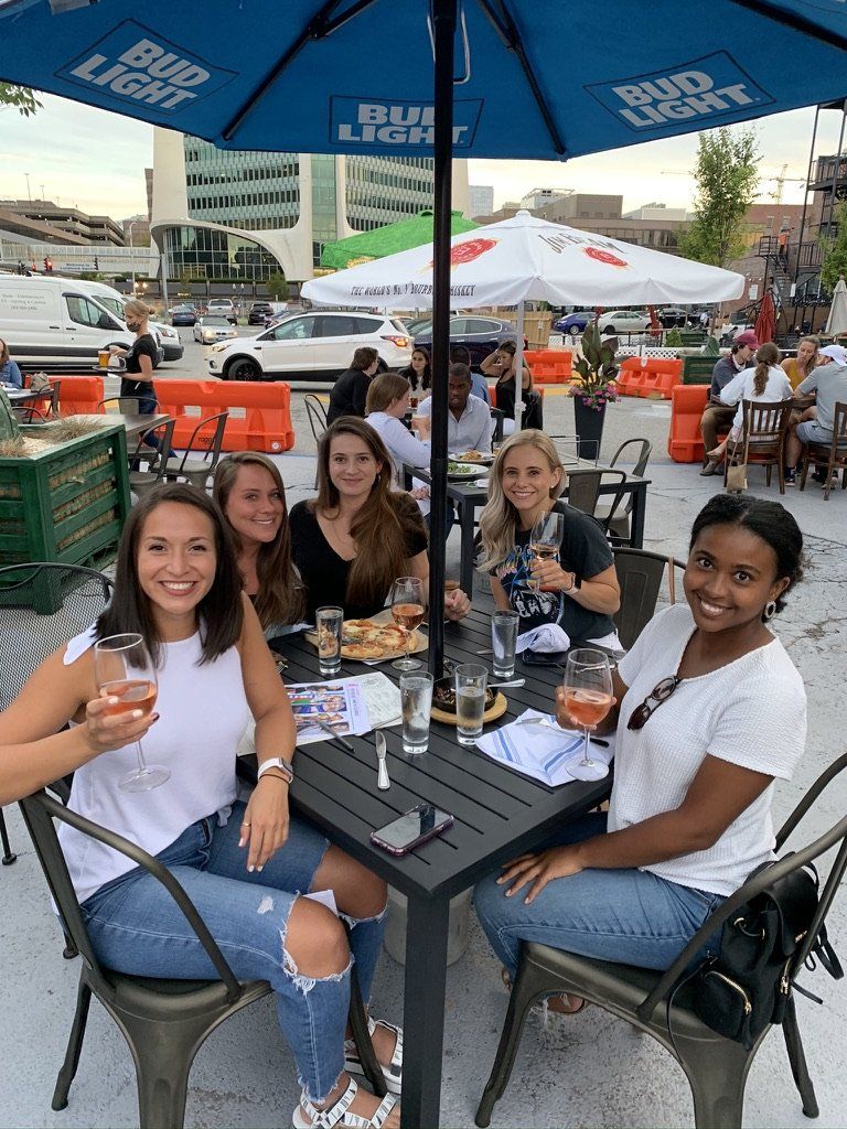 Group of people sit at outdoor table, drinks in hand, smiling. Umbrella overhead, city setting.