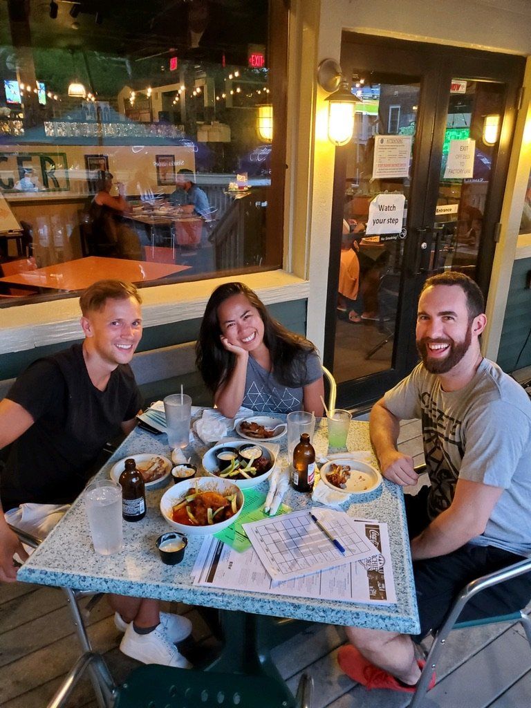 Three people smiling at a table with food and drinks outside a restaurant at night.