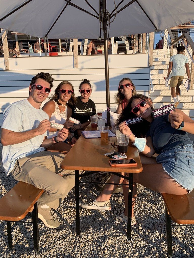 Group of people at outdoor table, smiling, holding items. Under umbrella, beach setting.