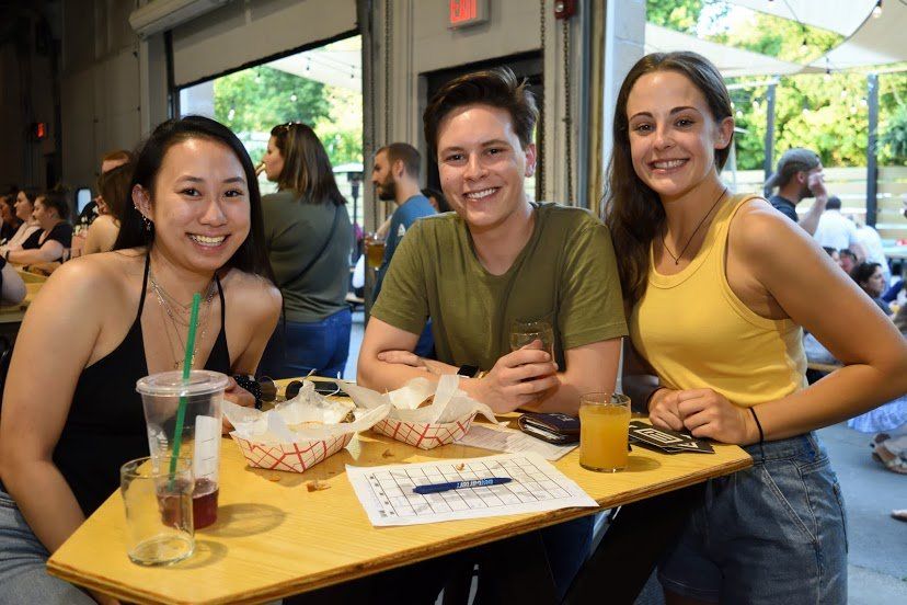 Three people smiling at a table with drinks and food at an outdoor venue.