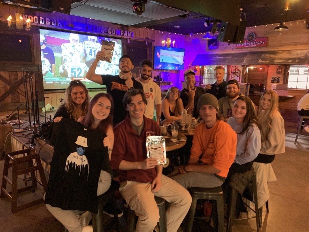 Group of people celebrating in a bar, holding awards. Large screen displaying a sporting event, wood interior.