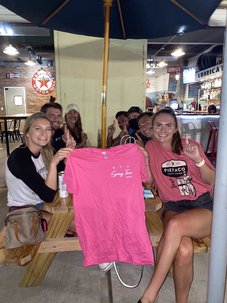 Group of people at a bar, holding up a pink shirt. People smile, some make hand gestures.