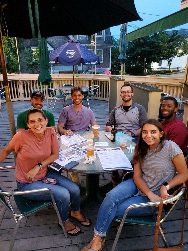 Group of people seated at an outdoor table under a patio umbrella, smiling and interacting.