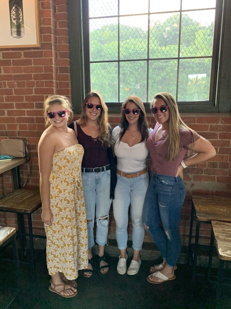Four women wearing sunglasses pose near a window; brick wall background.