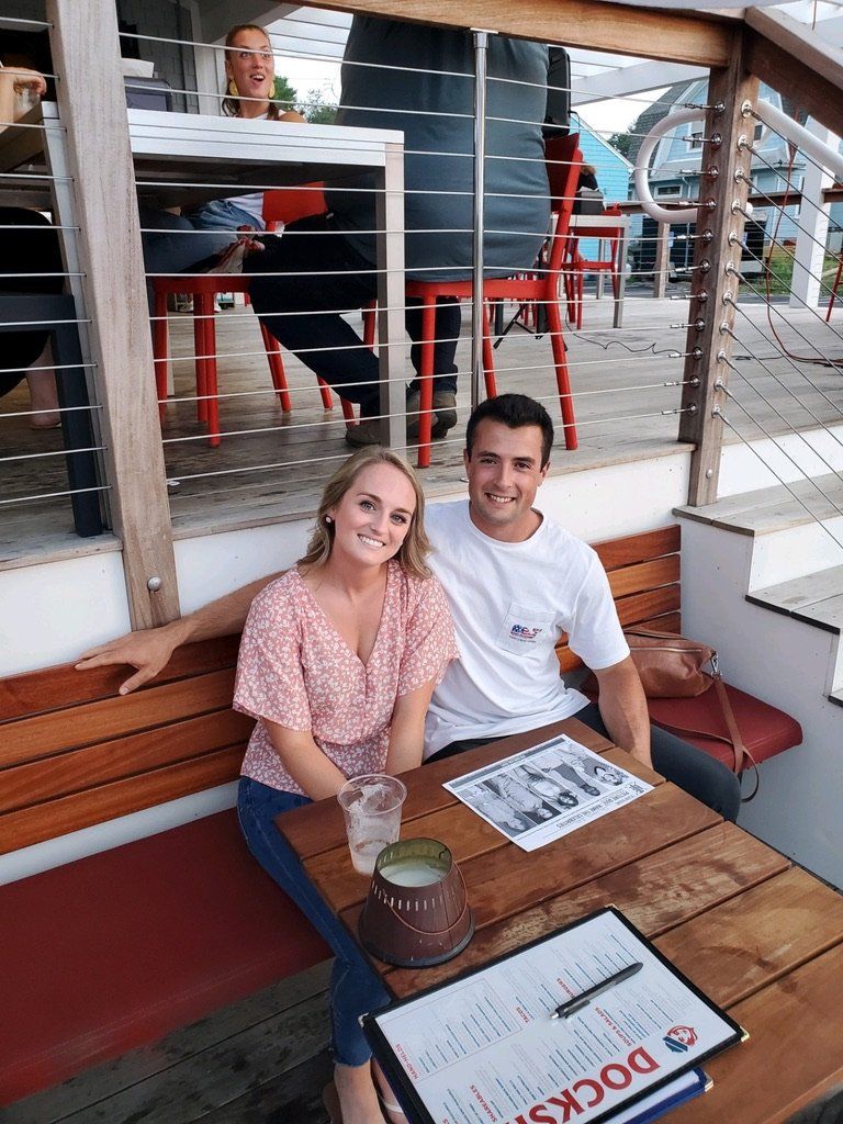 Couple seated at outdoor restaurant table; waterfront setting.