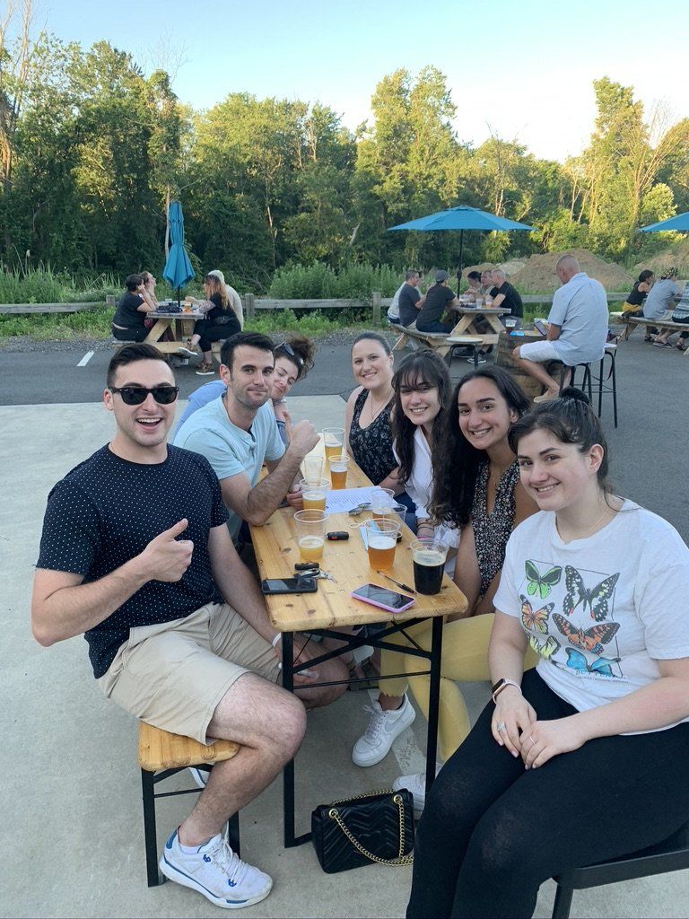 Group of people at an outdoor table; some with beers, smiling and looking at the camera. Trees and other tables in the background.