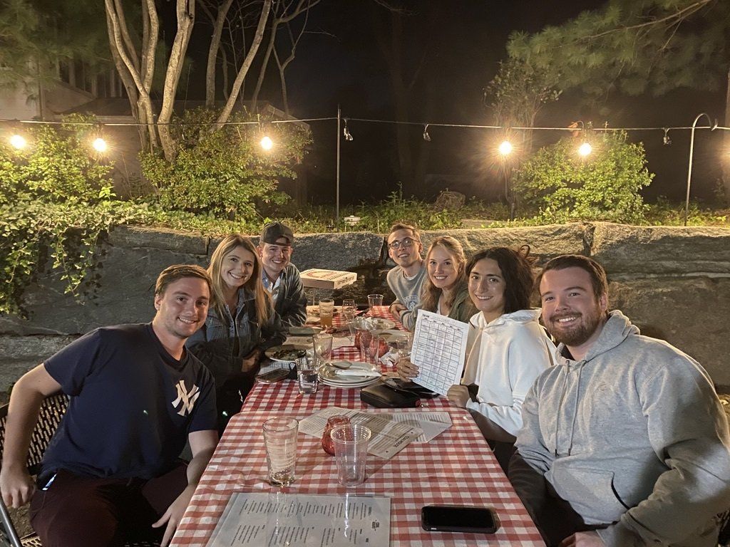 Group of friends smiling at an outdoor table with food, under string lights.