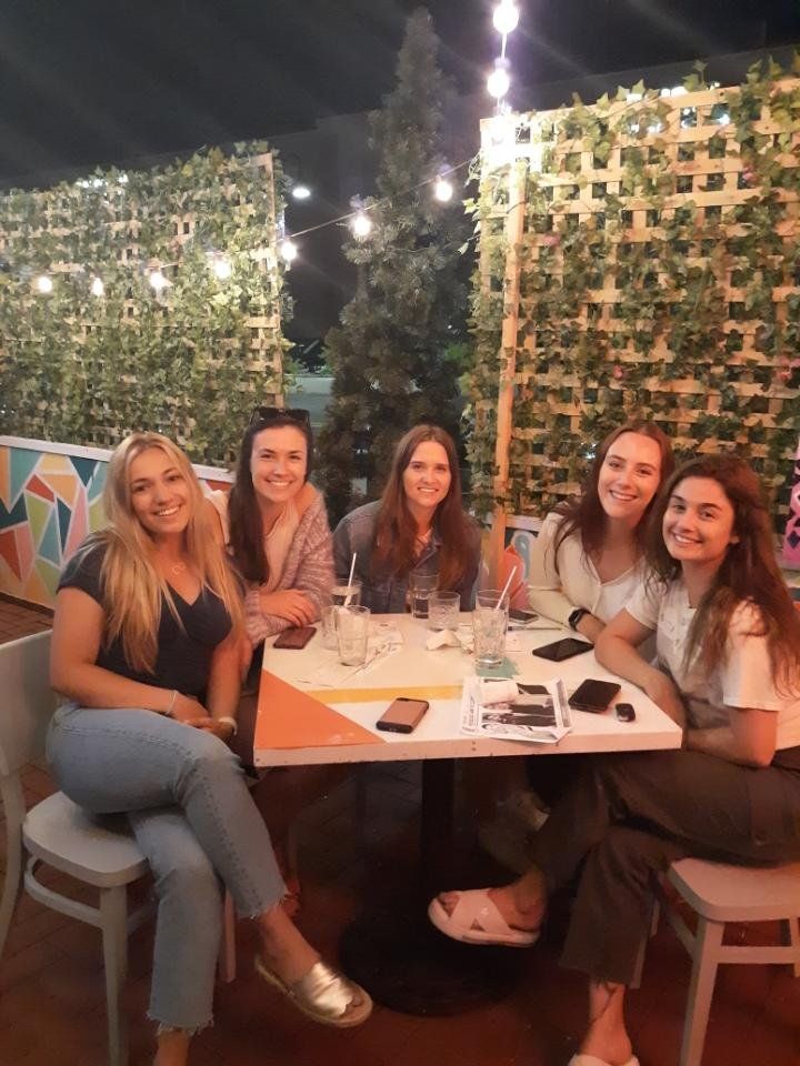 Five young women sit at an outdoor table, smiling at the camera. Twinkling lights and green wall in background.