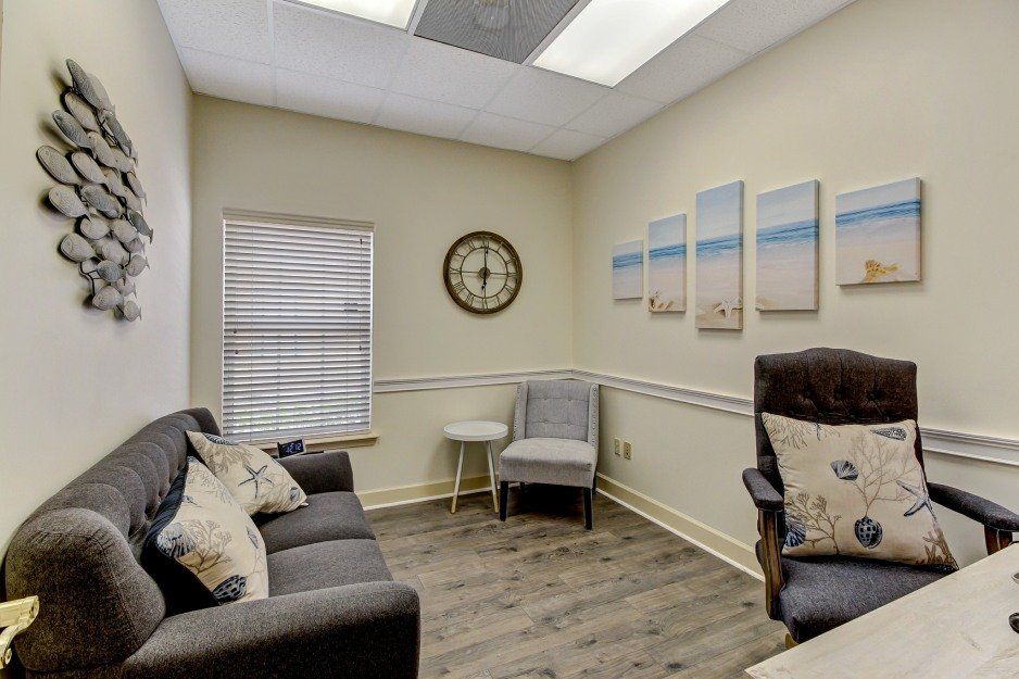 Small beige office waiting room with gray chairs, wall art, a clock, and a window blind