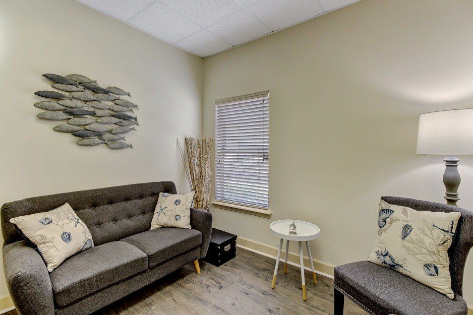 Small beige waiting room with gray sofa, wall art, side table, window blinds, and floor lamp