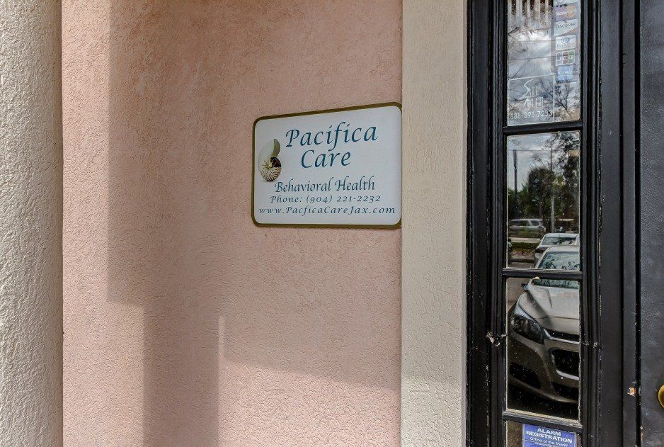 Pacifica Care sign on a beige wall beside a glass door with a parked car reflected nearby