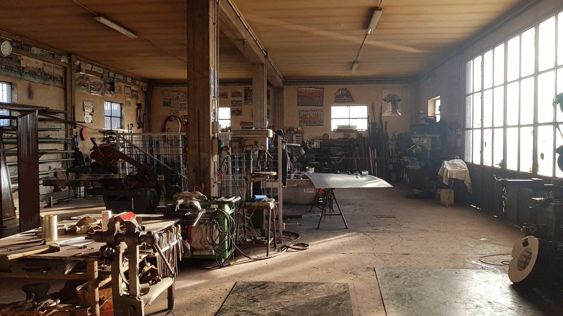 Interno di un'officina illuminata dal sole, con travi in legno, attrezzi e banchi da lavoro lungo ampie finestre.