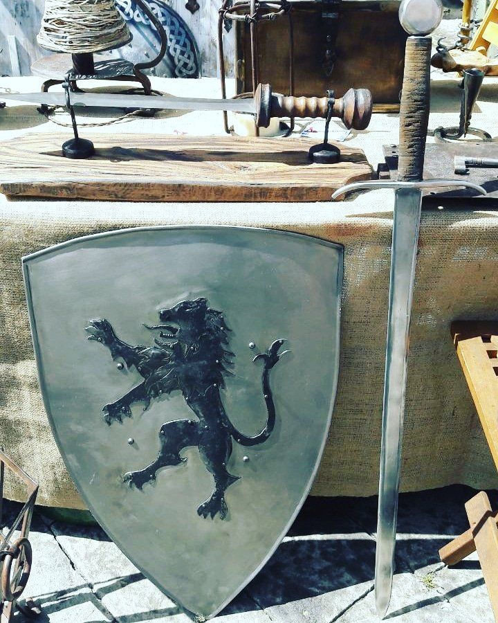 Scudo grigio con l'emblema di un leone nero appoggiato accanto a una lunga spada e armi di legno su una panca