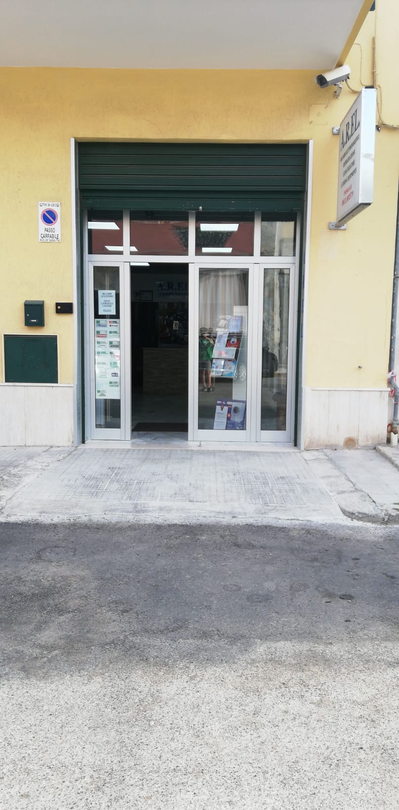 Ingresso dell'edificio con doppie porte a vetri, serranda verde e pareti gialle.