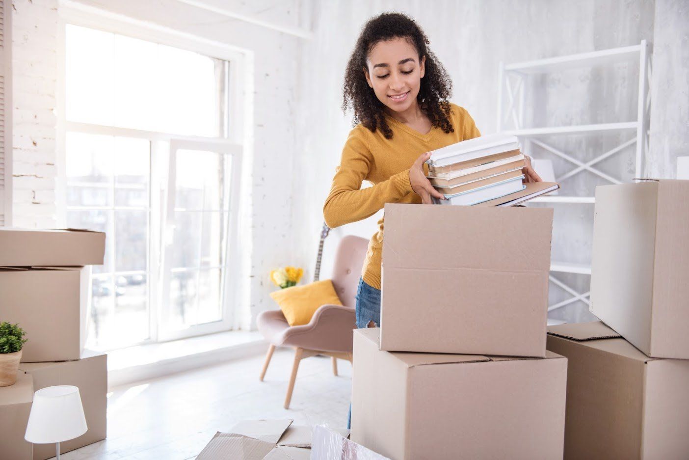 Woman in yellow shirt packing books into a cardboard box in a light-filled room.