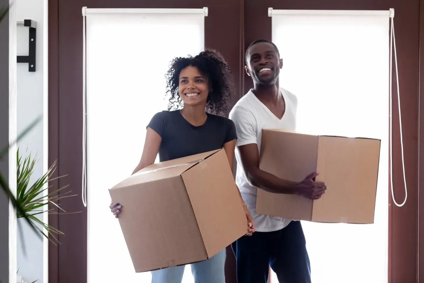 Couple, smiling, holding boxes, moving into a new home by a doorway.