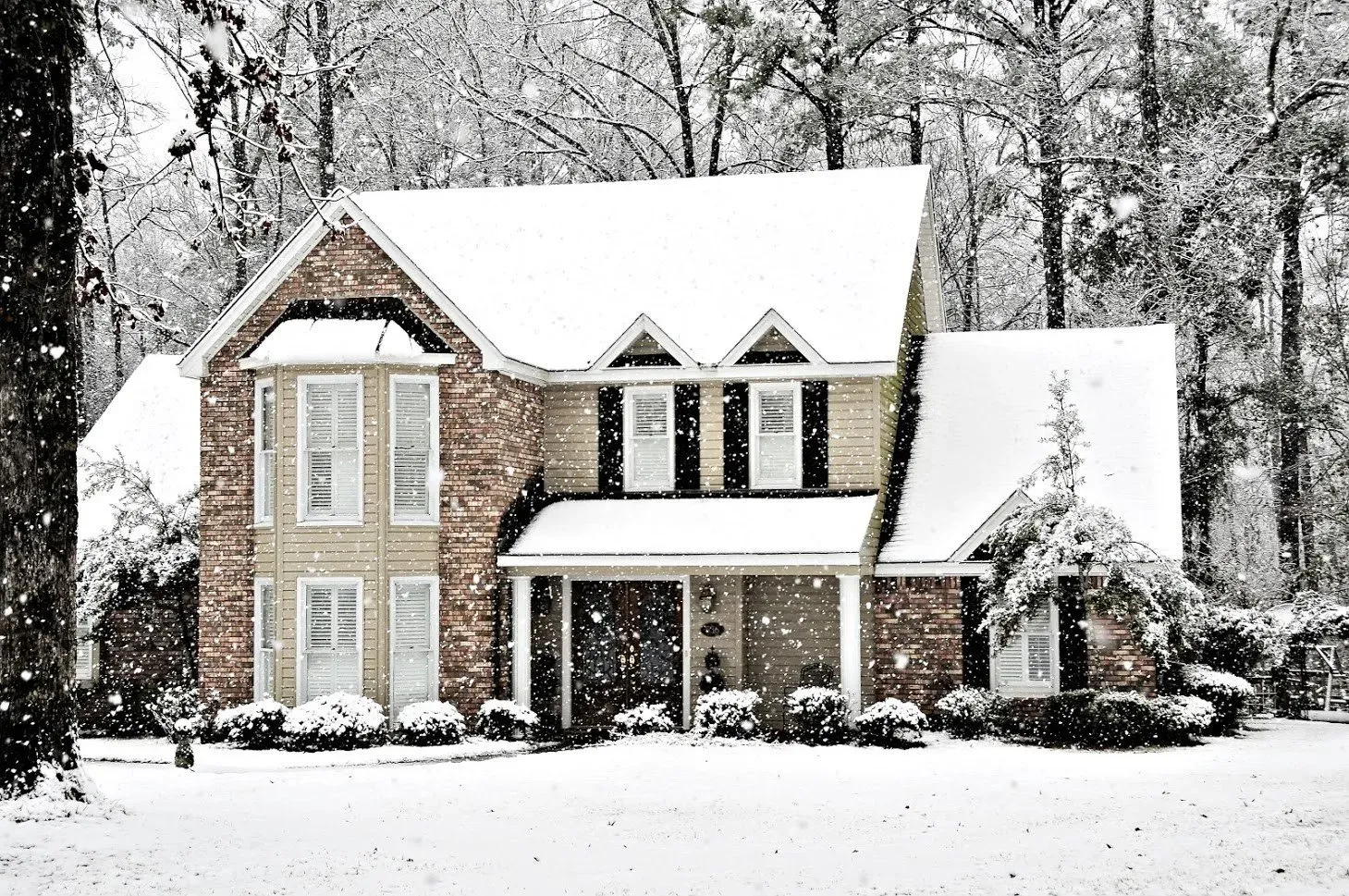 A two-story house covered in snow with a brick facade and trees in the background.