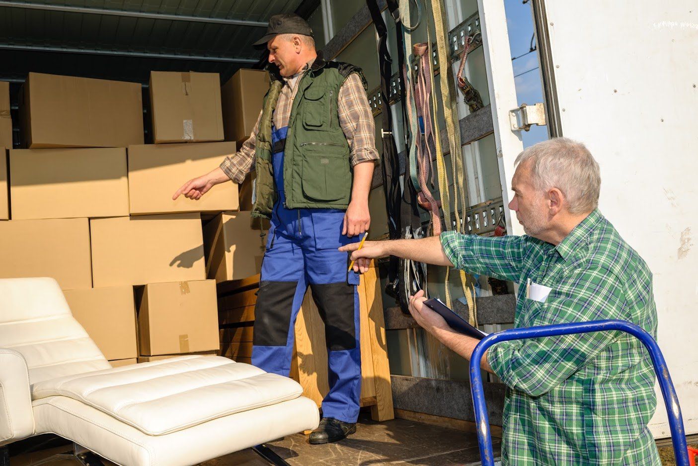 Two movers loading a truck: man in green vest and overalls points to boxes, other points with clipboard, white chaise visible.