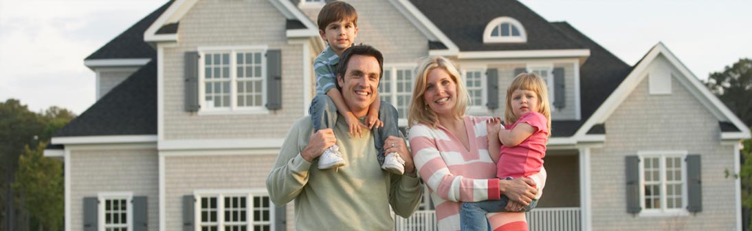 Family of four smiling in front of their new home. The dad has a boy on his shoulders and the mom holds a girl.