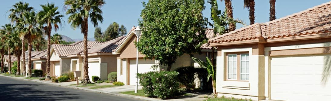 Row of houses with tan walls and red-tile roofs, palm trees, blue sky.
