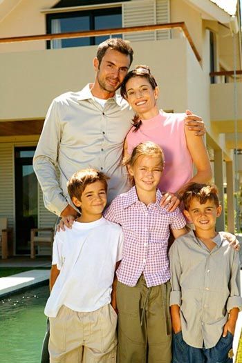 Family of five standing in front of a house, smiling; parents have arms around children.