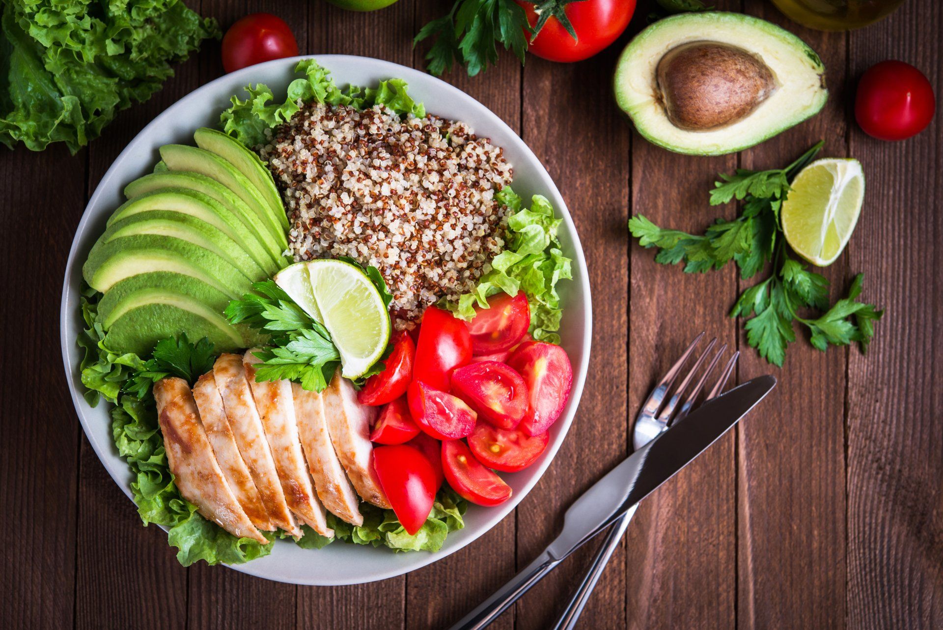 Healthy salad bowl with quinoa, tomatoes, chicken, avocado, lime and mixed greens, lettuce, parsley on wooden background top view. Food and health.