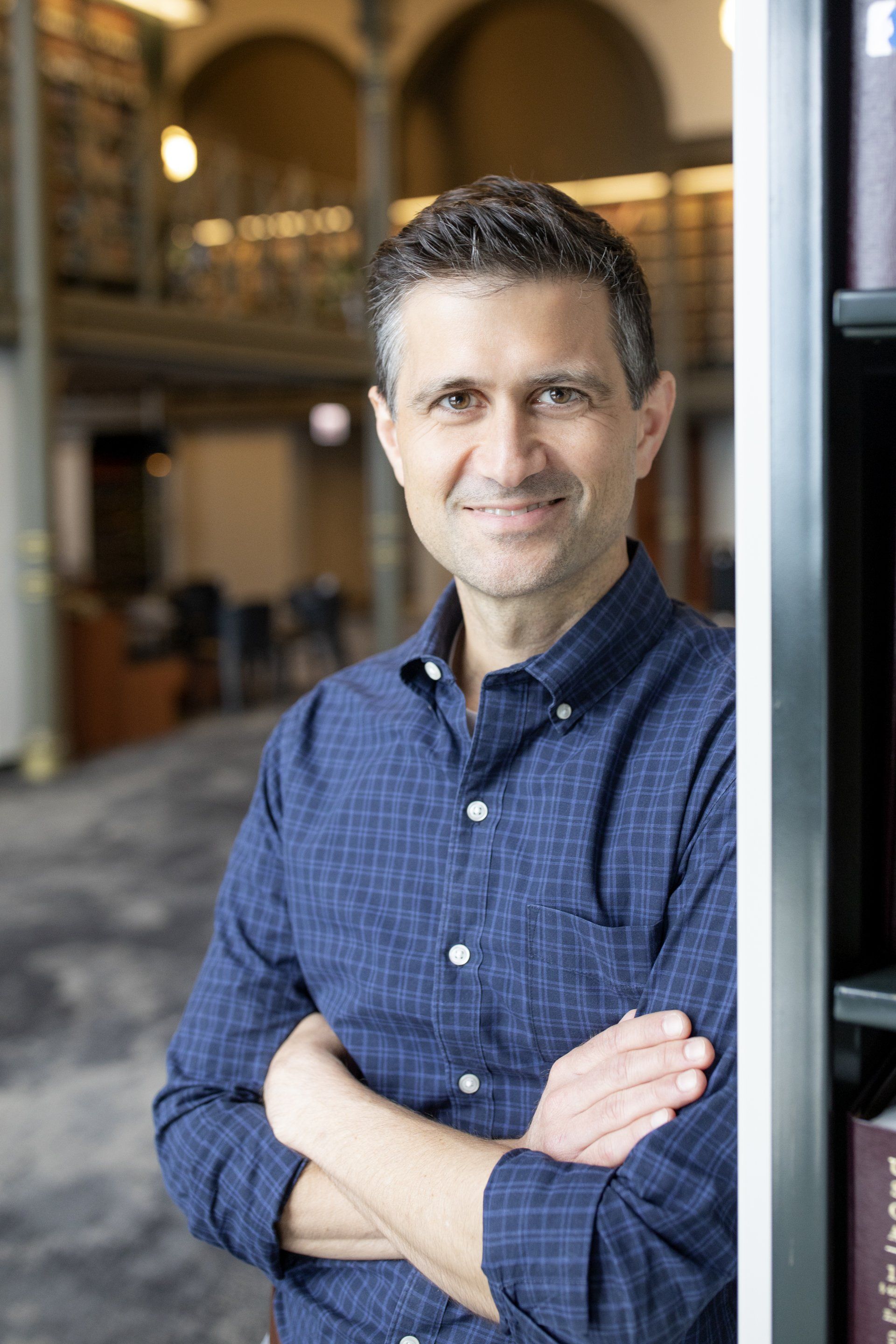 A man in a blue shirt is standing in front of a bookshelf with his arms crossed.