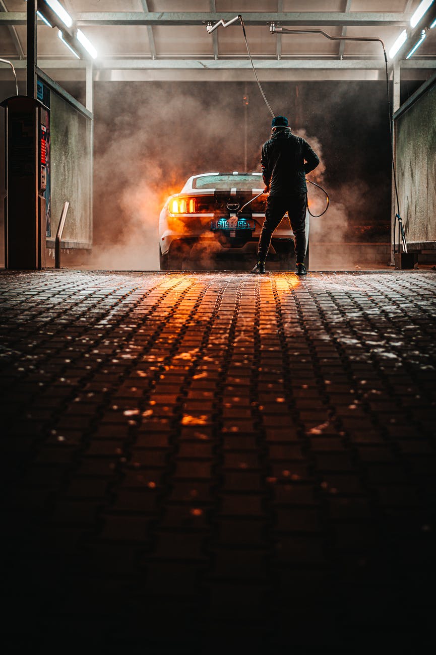 Person washes car at a car wash. The car and floor are illuminated in orange light; steam is rising.