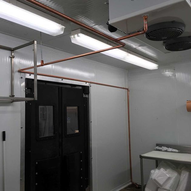 Interior of a walk-in cooler, featuring copper piping, fluorescent lights, a black door, and a table.