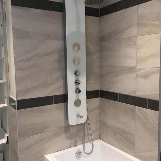Bathroom with a shower column and a small bathtub. Grey and white tiles with dark border.