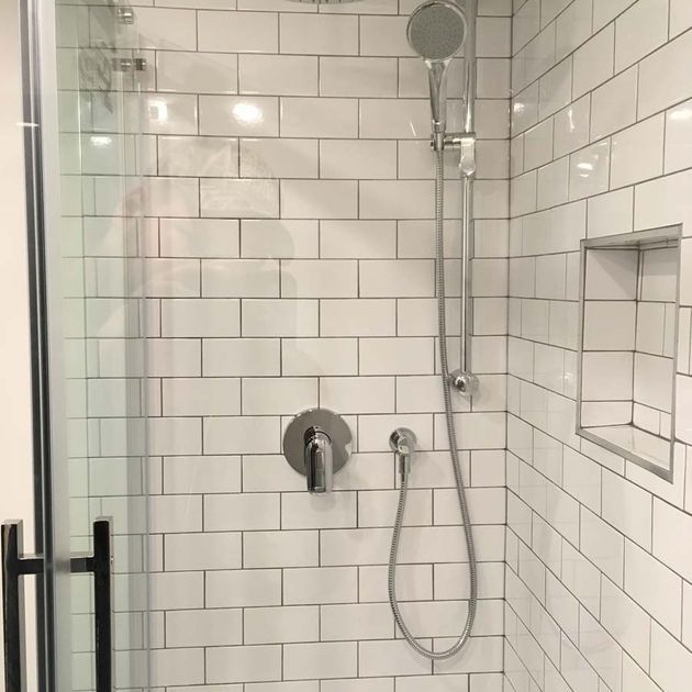 White-tiled shower with glass door. Chrome fixtures include a shower head, faucet, and recessed shelf.