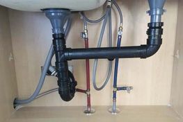 Under-sink plumbing: black drain pipes, silver supply lines, red and blue water lines in a wooden cabinet.