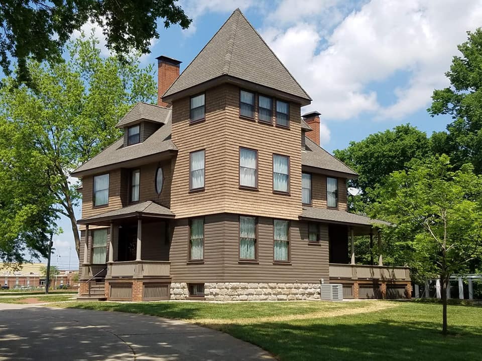 A large brown house with a gray roof is surrounded by trees and grass.