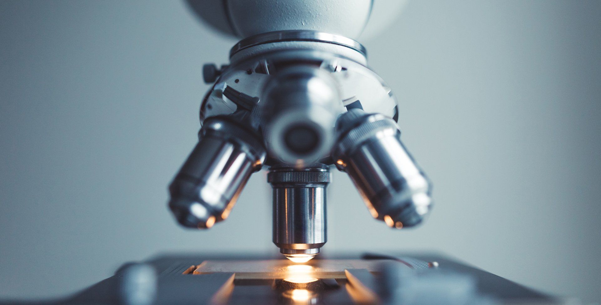 Close-up of a microscope with silver metal components, focused on the objective lens, against a soft gray background.