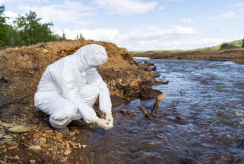 Person in white hazmat suit crouches at muddy river's edge, possibly collecting a water sample.