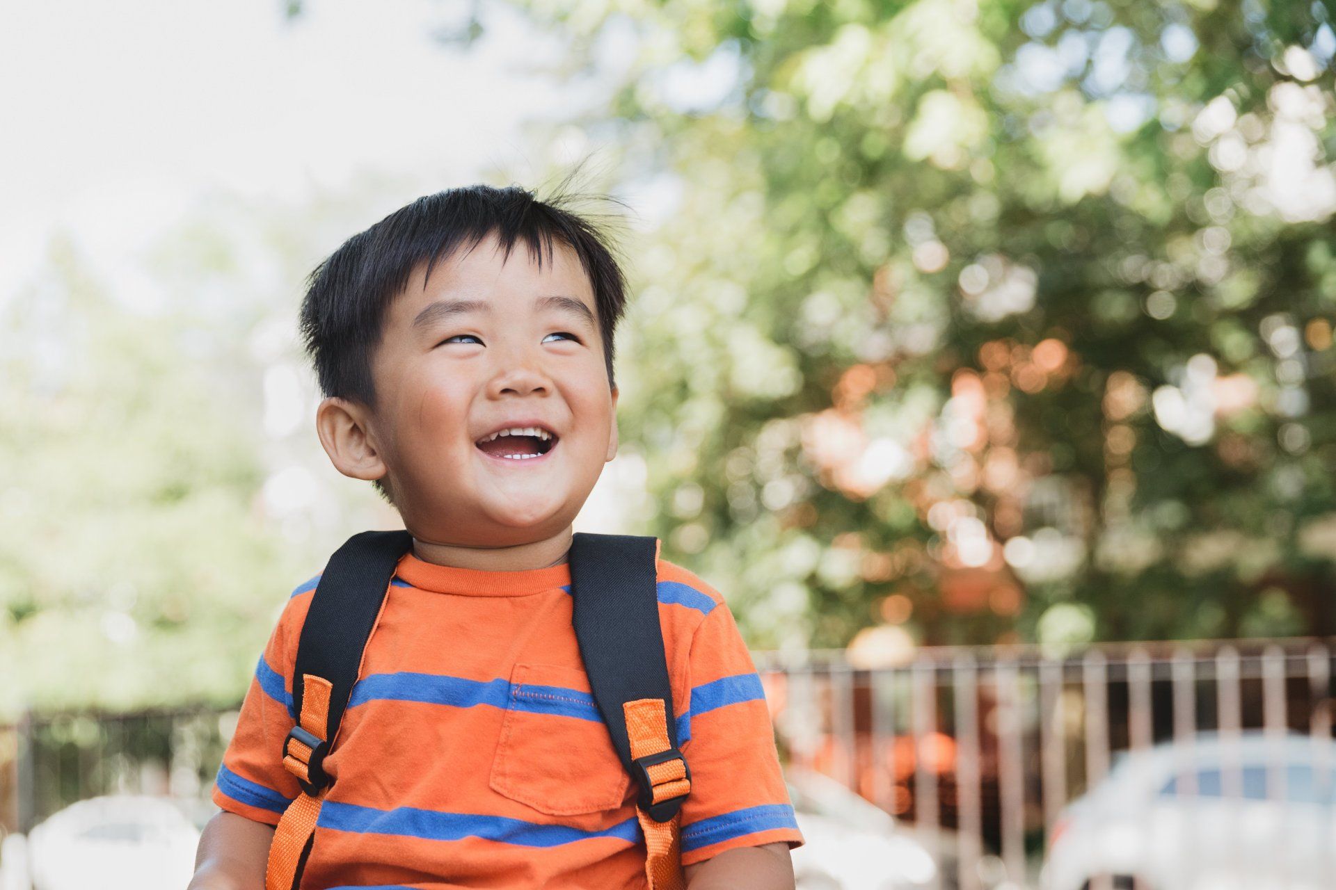 A young boy with a backpack is sitting in a park and smiling.