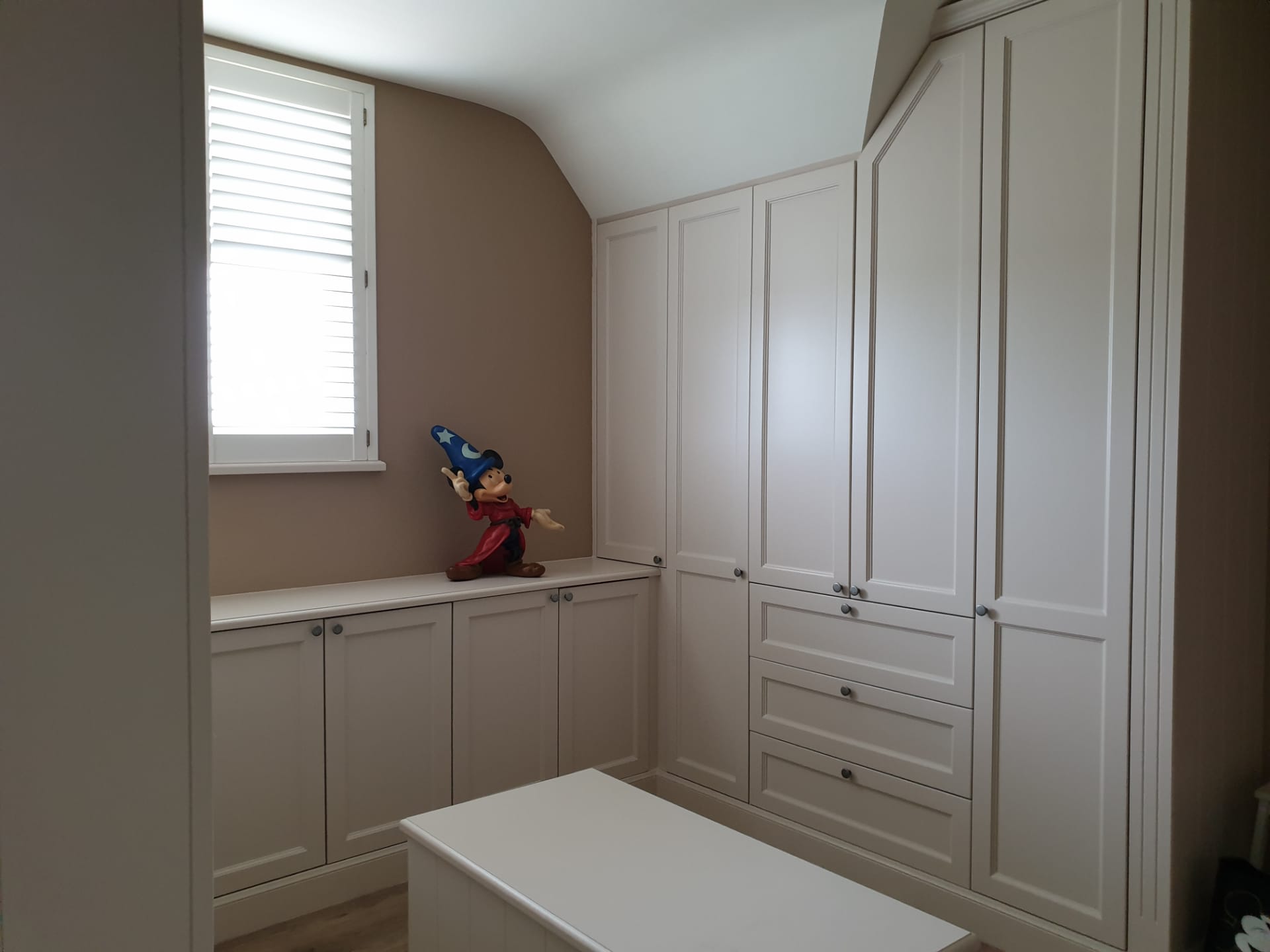 Houten dressing in landelijke stijl. Slaapkamer met ingebouwde crèmekleurige kasten, een raam en een beeld van een tovenaar.