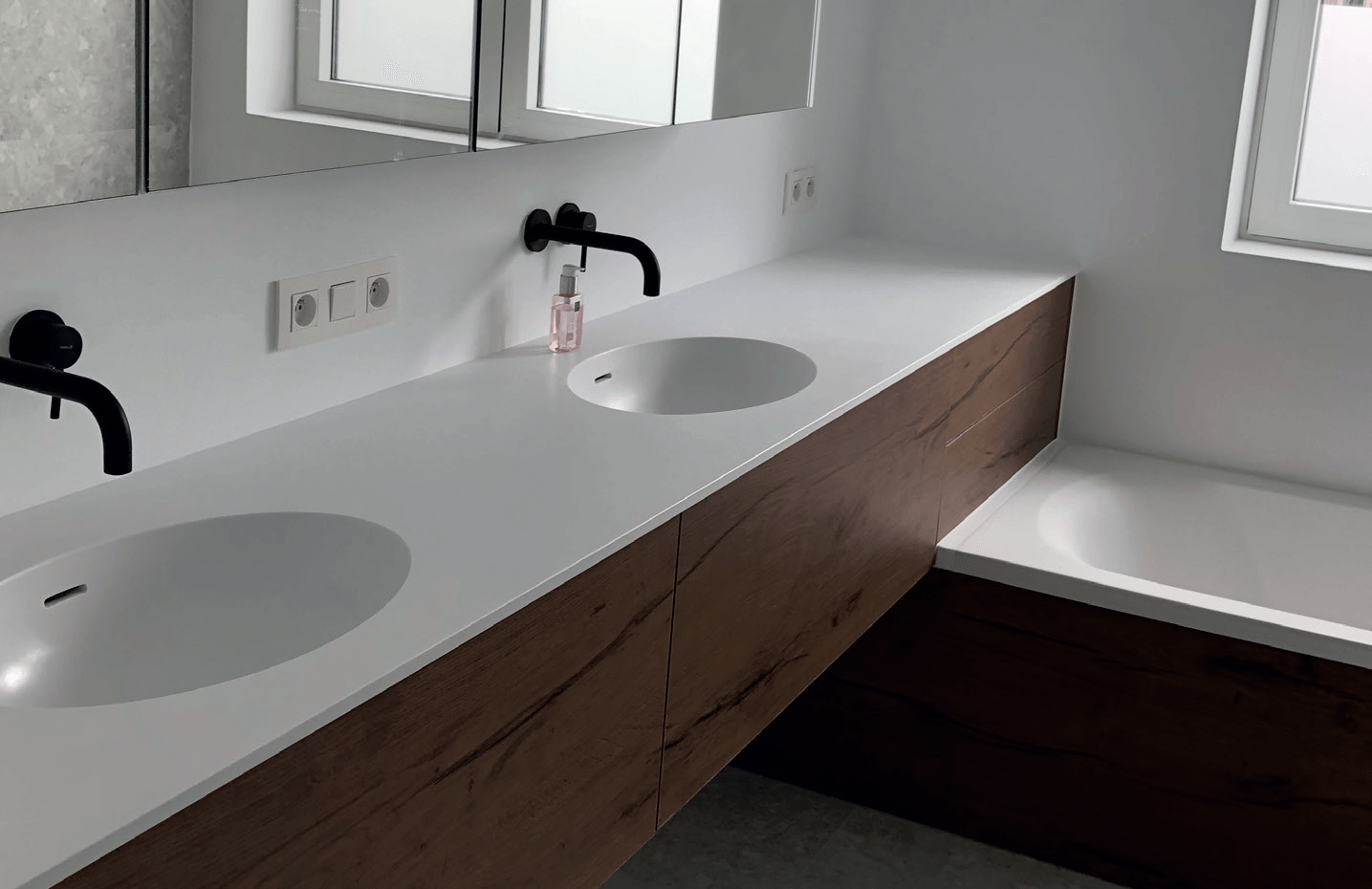 Moderne badkamer met een wit aanrechtblad, twee wastafels en een houten kast. Zwarte kranen en een badkuip.